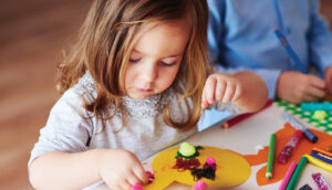 A young girl uses her right hand to place a variety of small, colorful objects onto a piece of yellow construction paper cut into the shape of a rabbit head.
